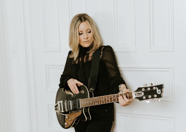 Bandfoto Christina Martin: Eine Musikerin mit langen blonden Haaren steht vor einer weißen Wand mit Kassettenstruktur. Sie trägt ein schwarzes Outfit und hält eine elektrische Gitarre in den Händen. Ihr Blick ist nach unten auf das Instrument gerichtet, w